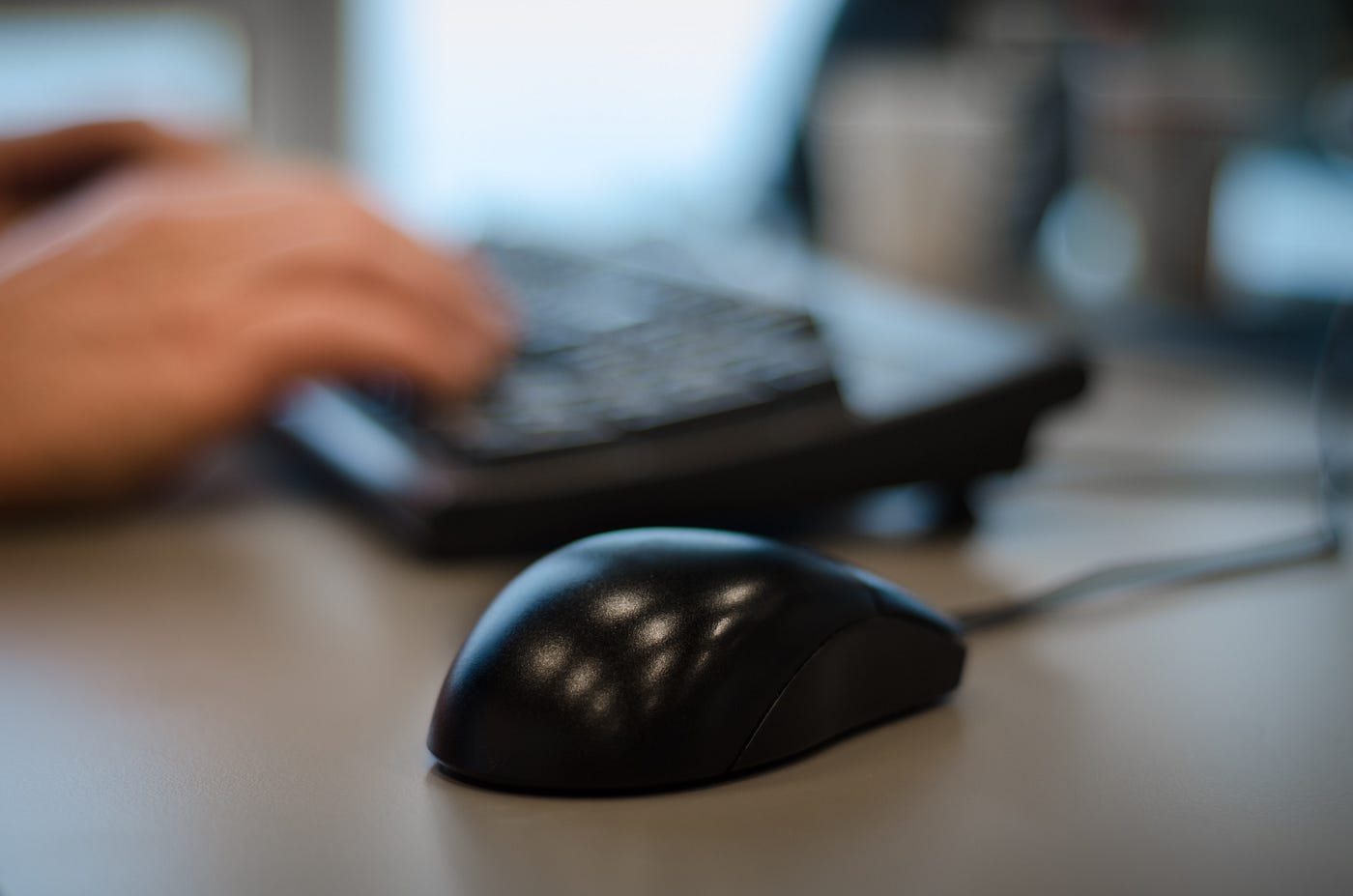 A computer mouse on a desk with hands on a keyboard slightly blurred in the background.