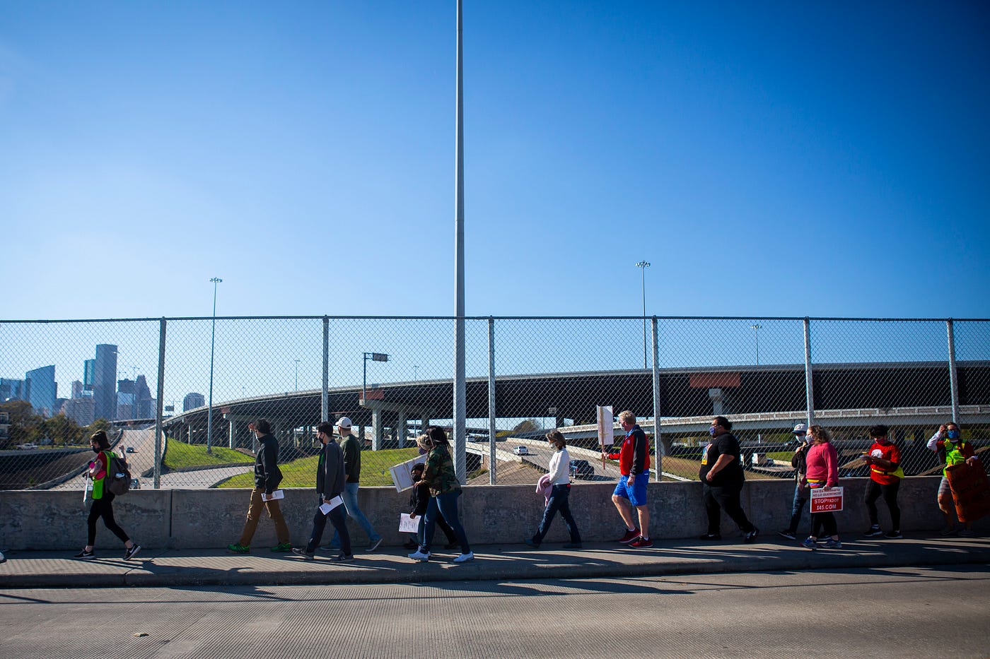 People carrying signs walk on a narrow sidewalk along a freeway. Behind them is a chain-link fence, and more freeways and a skyline in the distance.