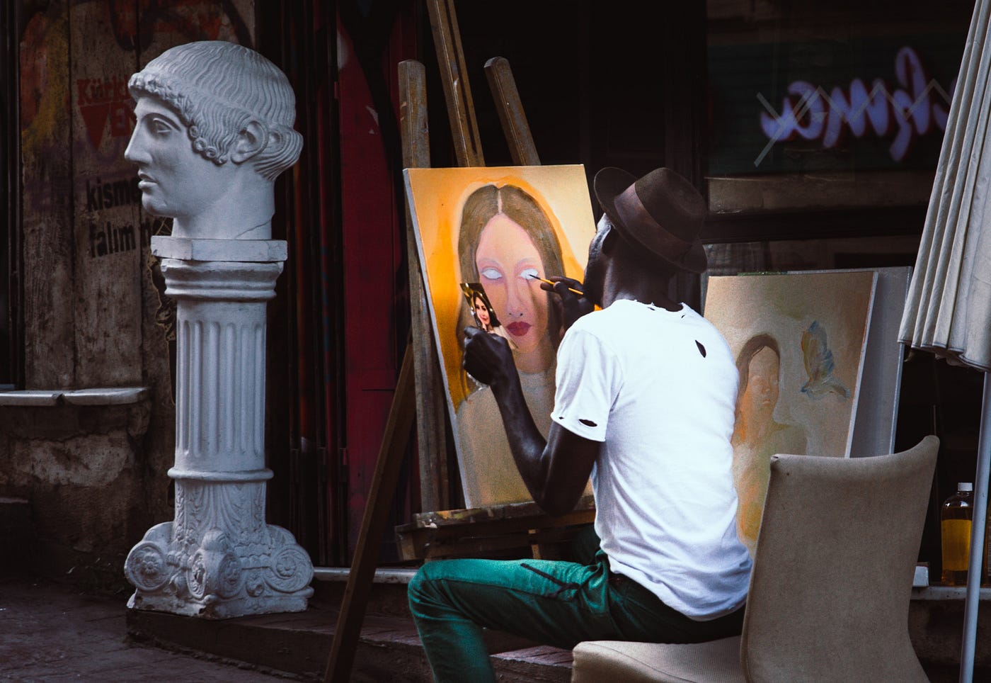 A Black man sits in a room painting a portrait of a woman. On the side of the room is a statue with a bust of a man on top of it.
