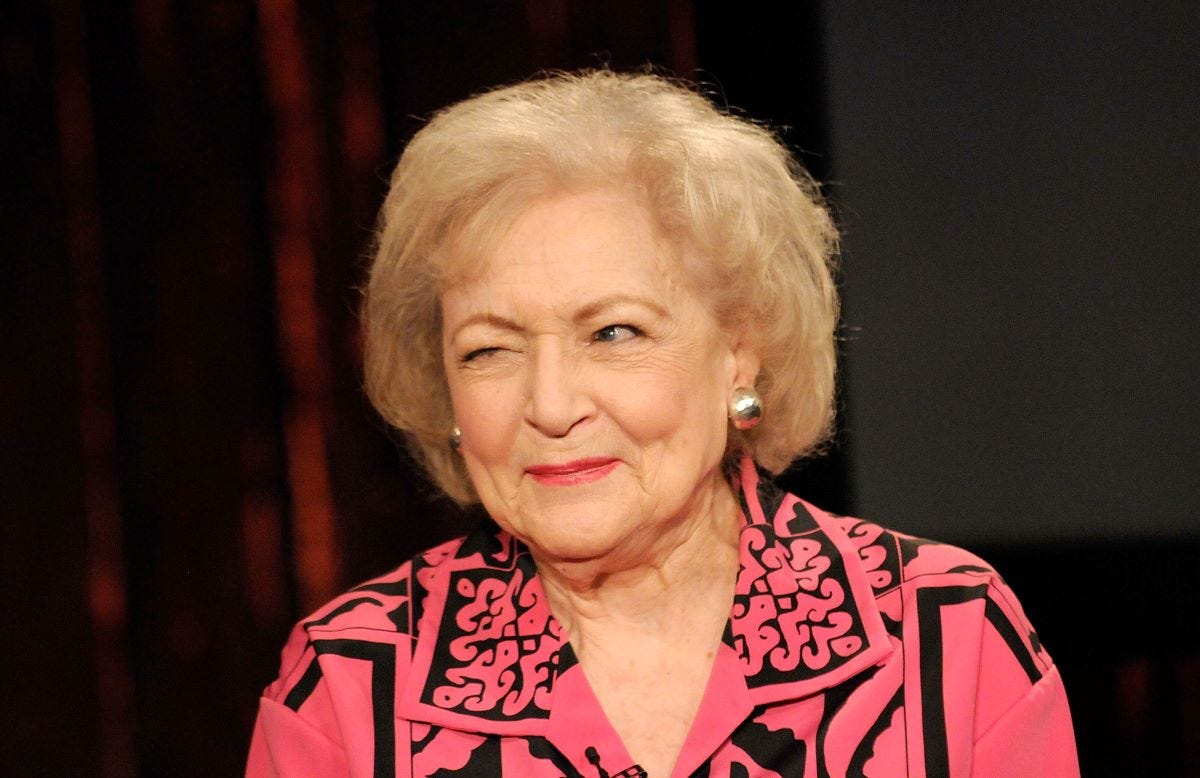 Betty White in a pinkish salmon colored blouse with black line detailing, winking off camera
