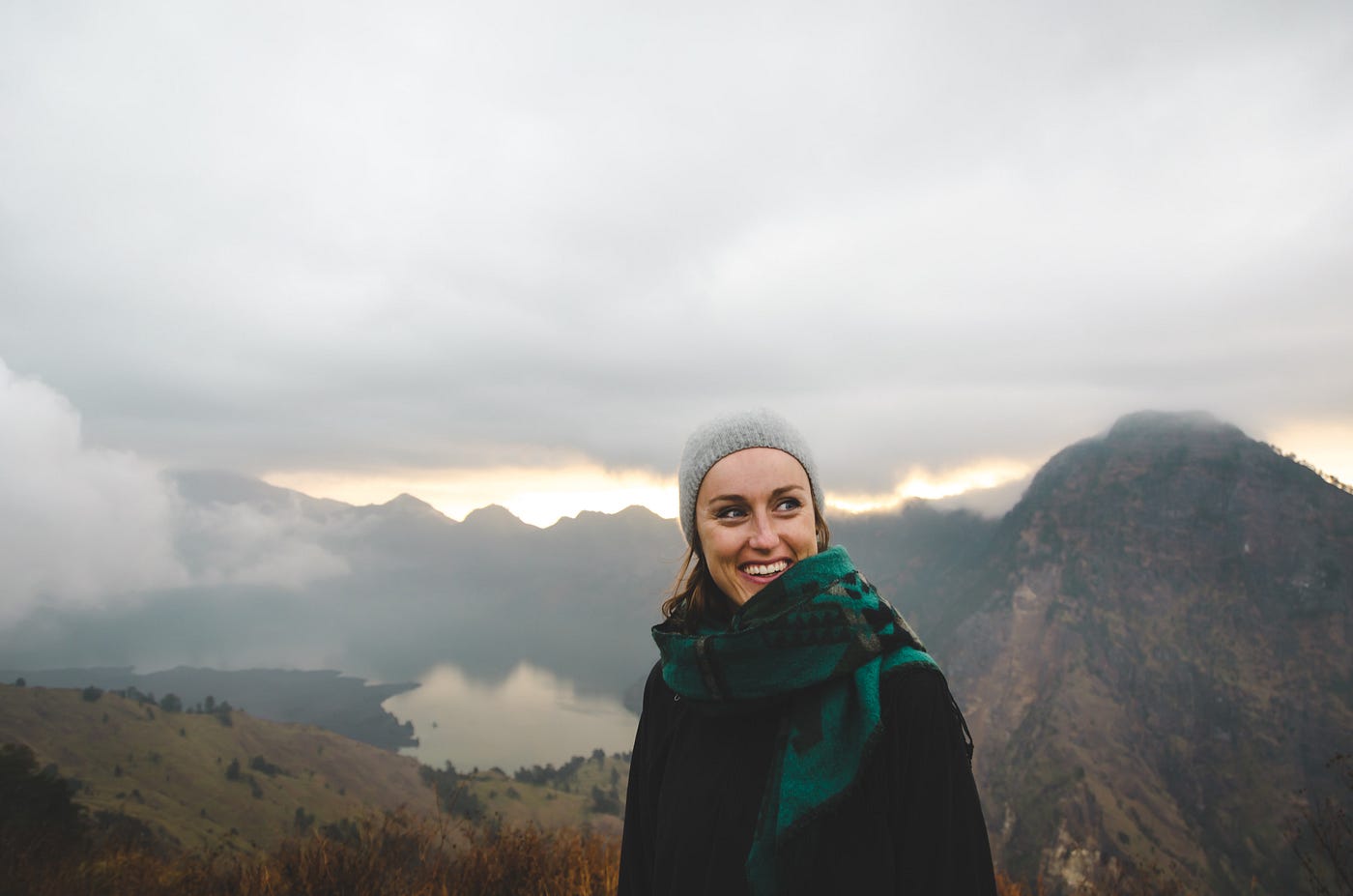 Photo of a woman in scarf, coat and hat smiling with a scenic background of hills and lake.