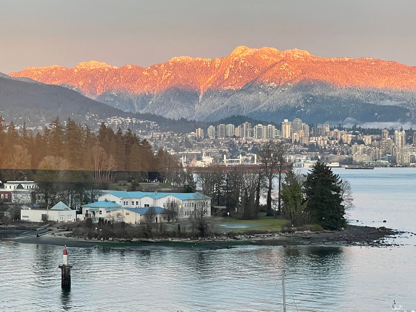 Rosy late light illuminating snow covered mountains behind sky scrappers. A harbour island with evergreen forest, mists