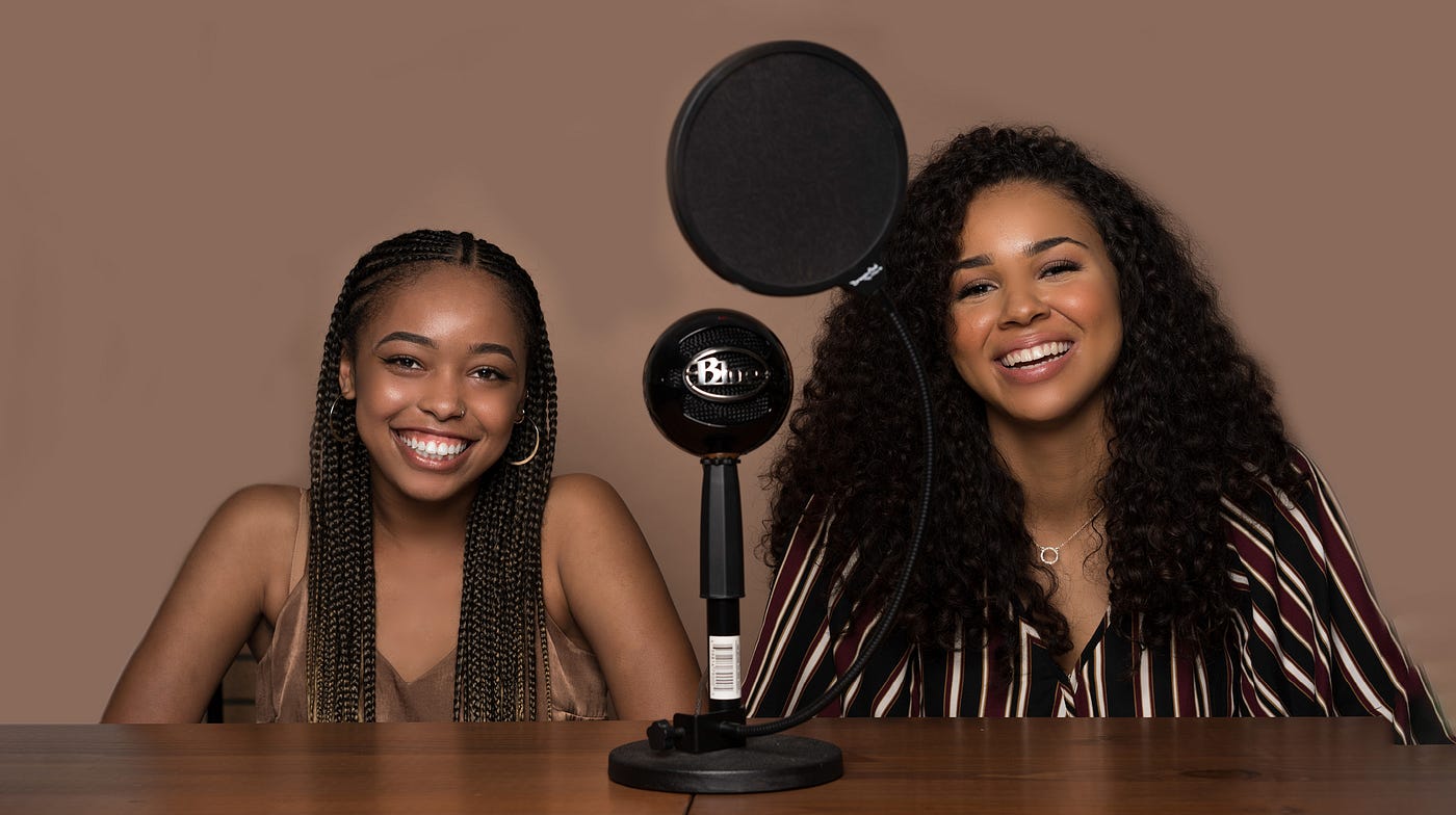 Sydney Charles and Tatum Larsen sit behind a wooden table smiling. On the table between the two of them is a microphone.