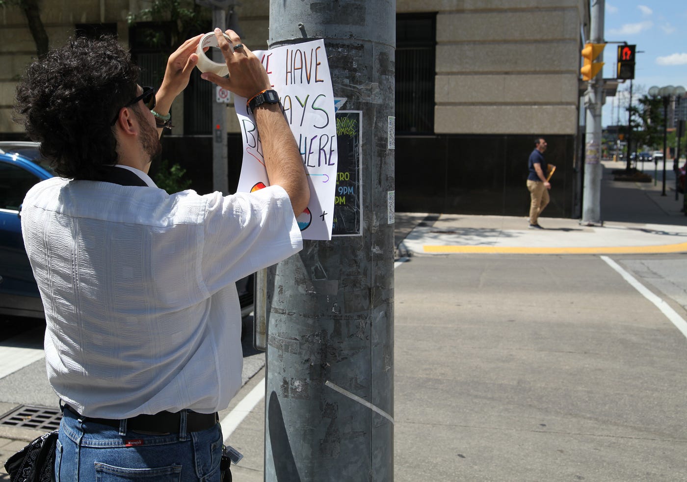 Michael tapes an activism poster that says “We have always been here” to a pole in Downtown Windsor.