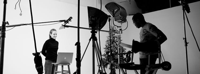 Woman with a laptop stands on a small production set, with a man operating a video camera.