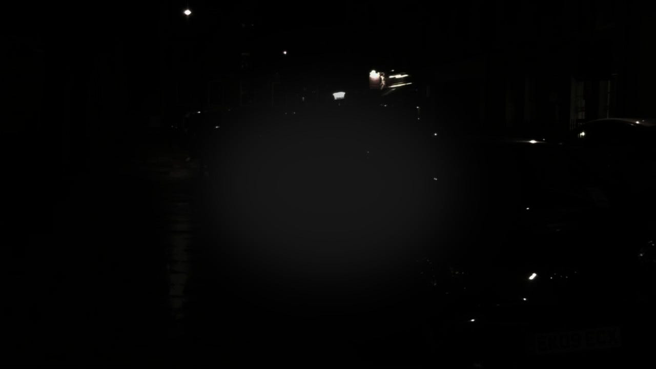 Mock-up of night blindness with central vision loss. There’s a large patch of grey in the center, blocking view of a line of cars along the street at night. Almost everything is pitch black save the glow from lights and reflections off car surfaces.