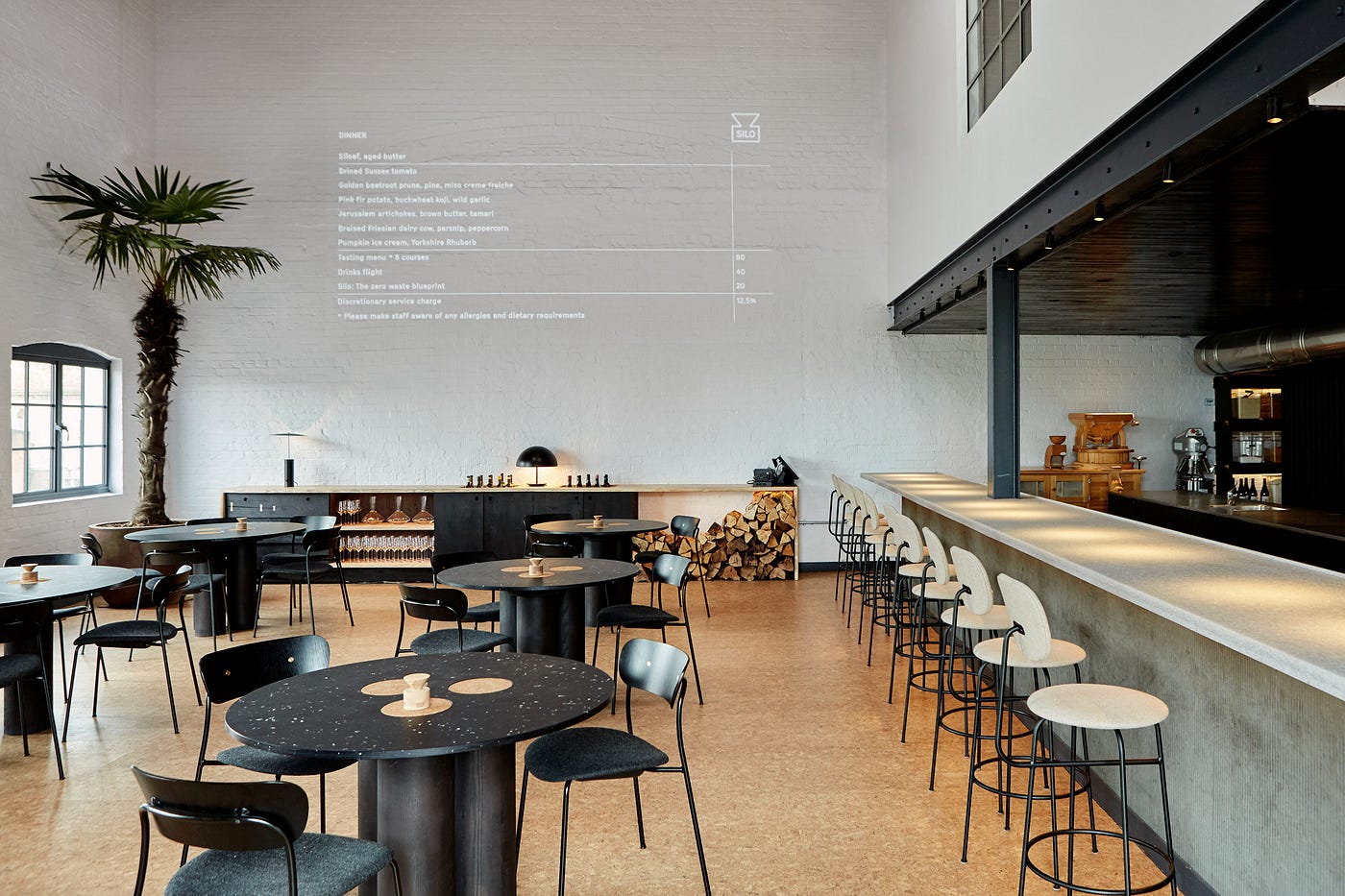I a room, text is projected on the wall, in front of it there are tables with chairs and on the right side is a long bar.