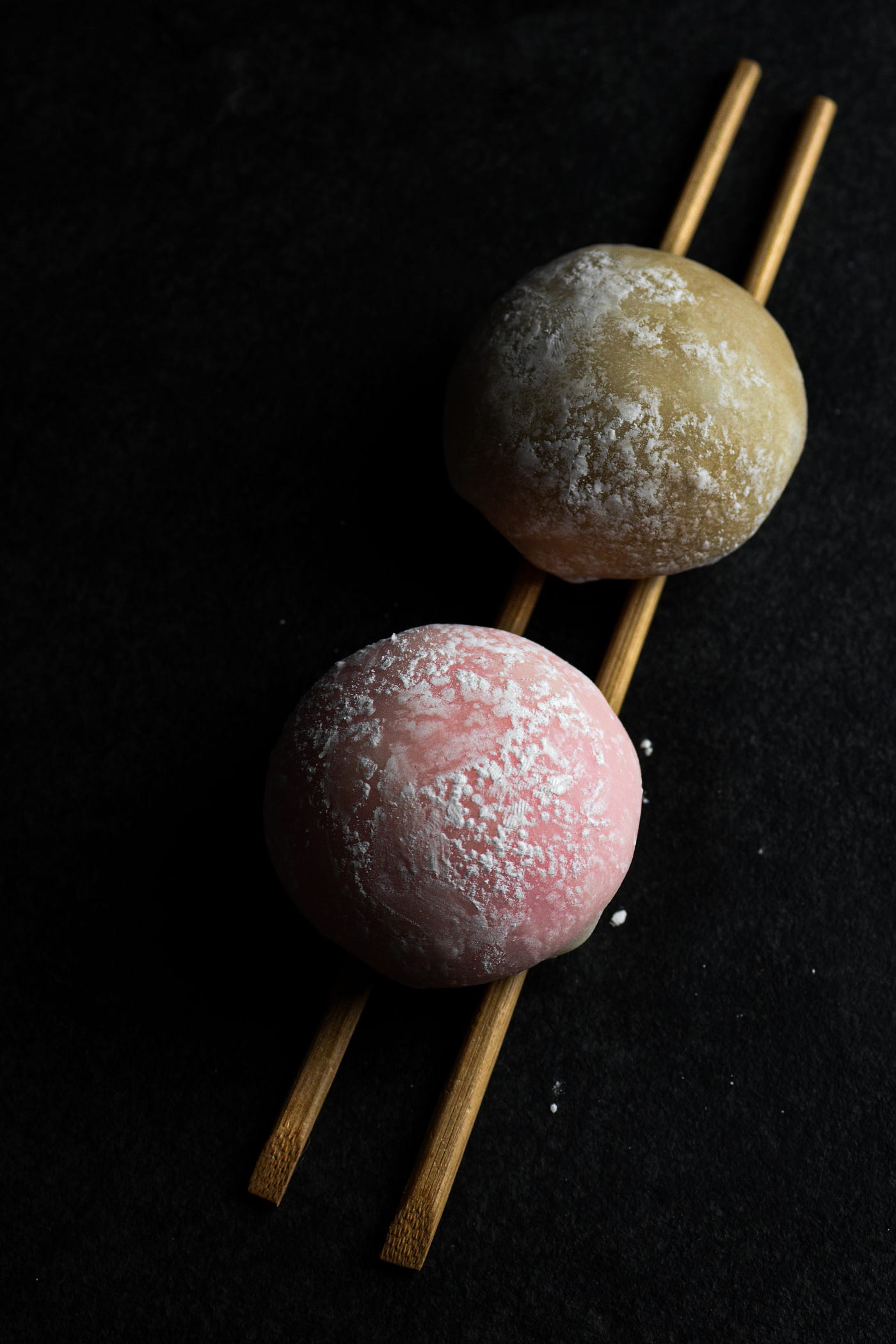 Two Mochi balls, one green one pink balanceing on top of a pair of chopsticks surounded by darkness.