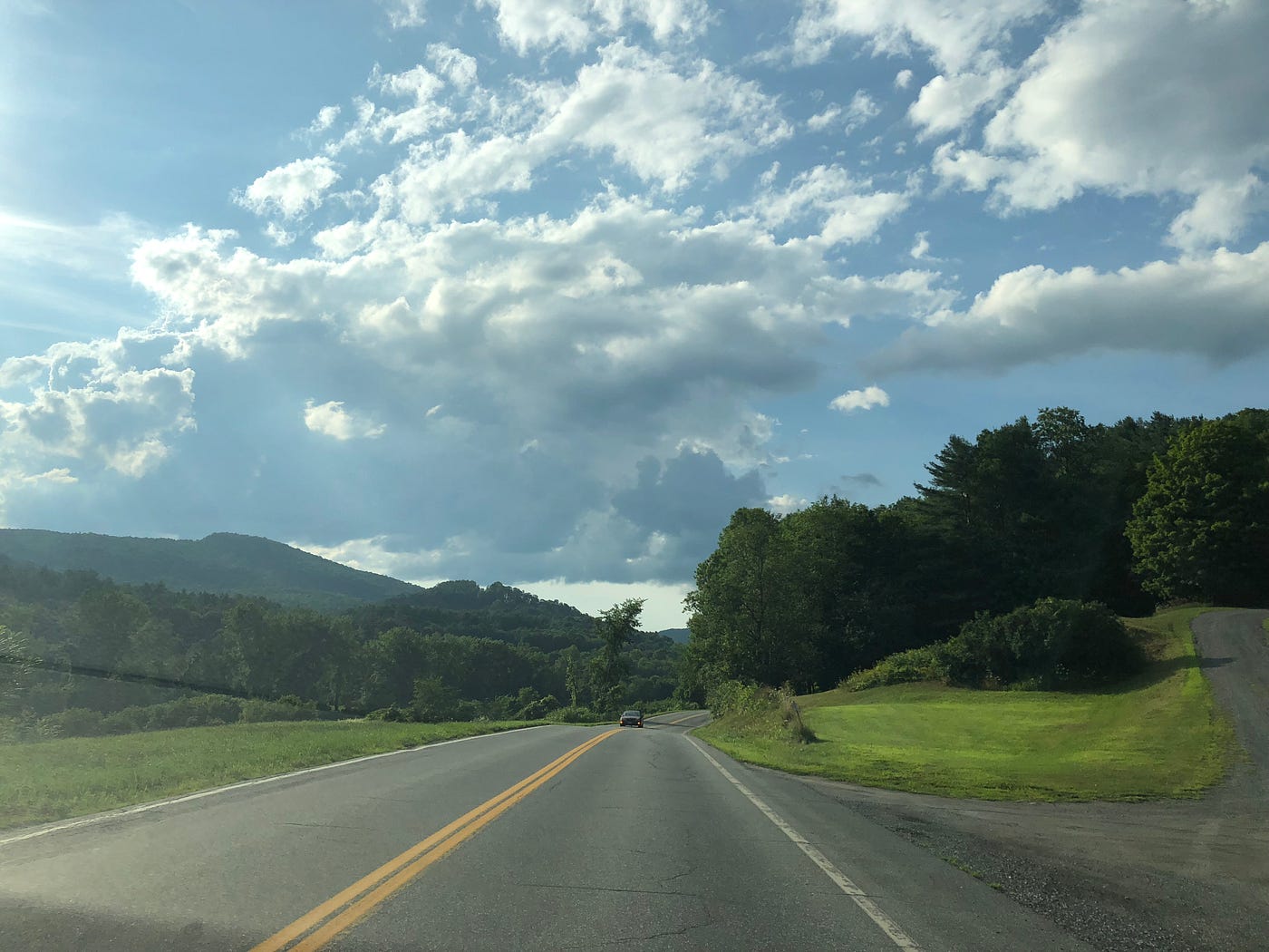 A highway in Vermont, lined with trees, and white clouds and mountains in the distance against a blue sky.