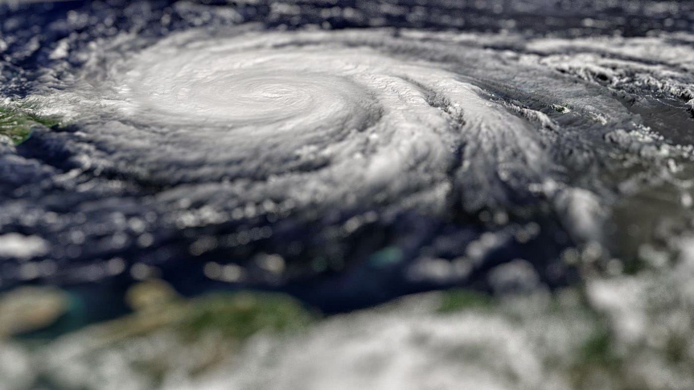 An angled aerial view photo of Hurricane Maria. The hurricane is seen as a large and tightly knit swirl of clouds. The surrounding deep blue sky and land beneath is blurred.