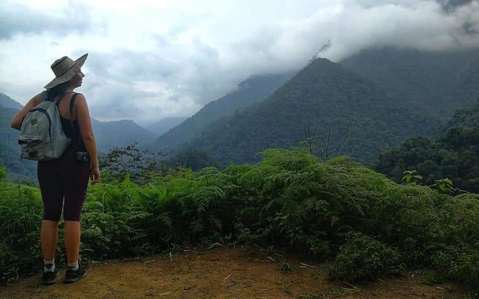 Kate Hillier stood with a backpack on, stood on the edge of a rainforest, looking at mountains covered in trees with clouds across the peaks