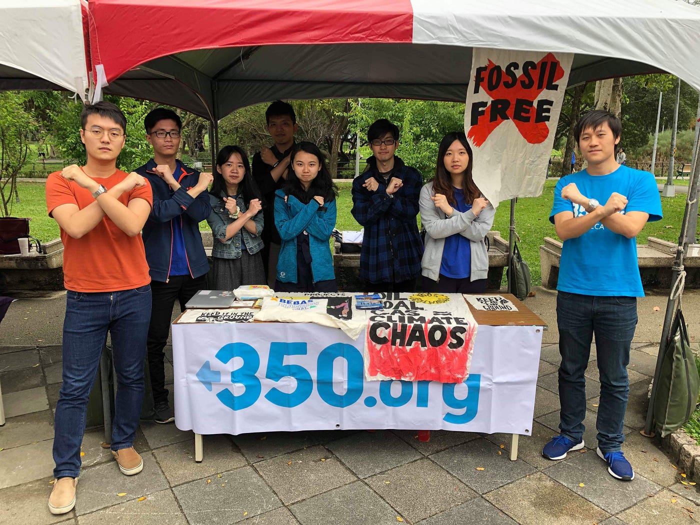 A group of people make Xs across their chests and stand under a tent around a table with a hanging poster reading “350.org.” A painted banner hangs off the table and reads “Oil, Coal, Gas = Climate Chaos.” Another poster hangs behind the people, reading “Fossil Free” with a red X behind it.