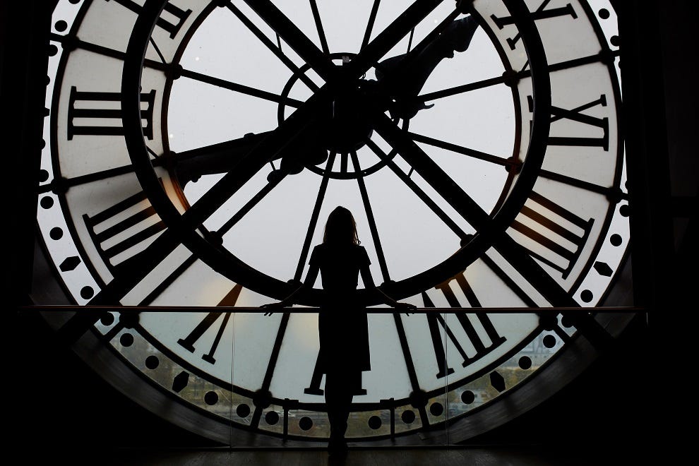 The silhouette of a tall woman stands at a large portrait window facing a clock