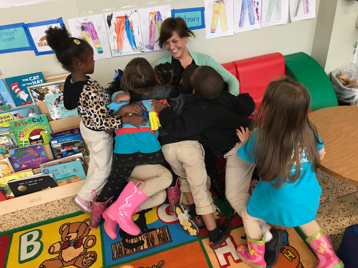 A smiling teacher sitting on a small couch under a pile of students who are hugging them.