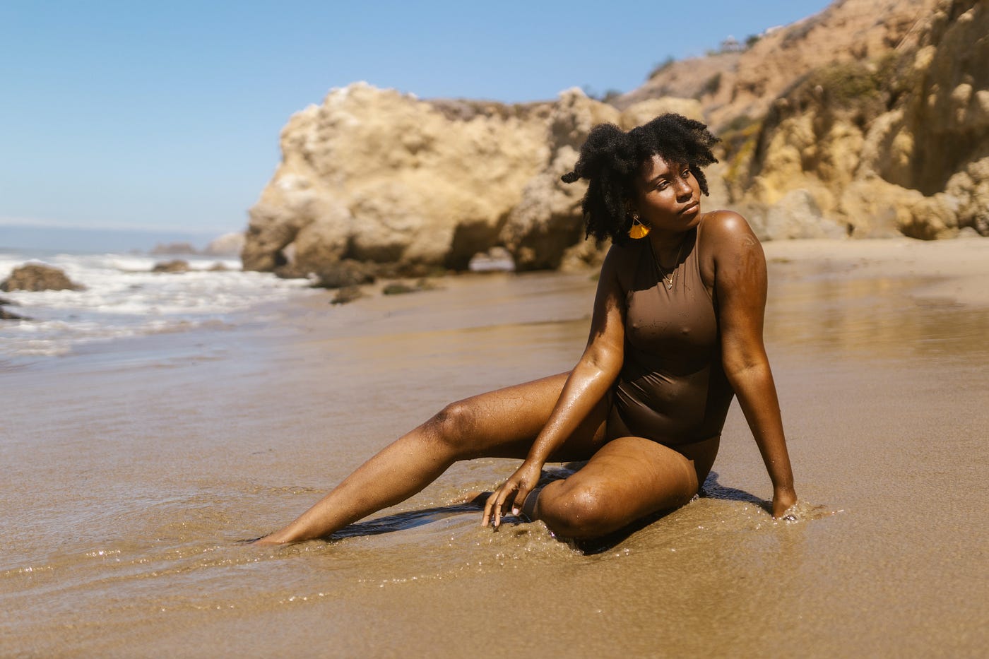 A Black woman lies on a sandy beach with a brown swimsuit on.