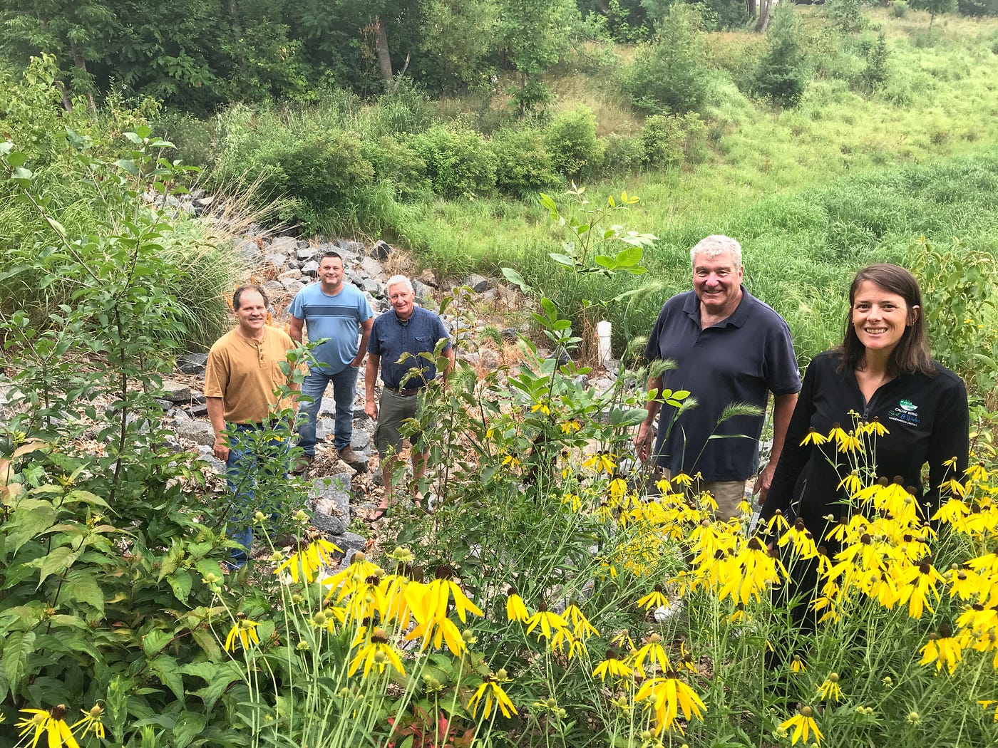 Five people stand in a spot with yellow flowers in the foreground and trees in the background.