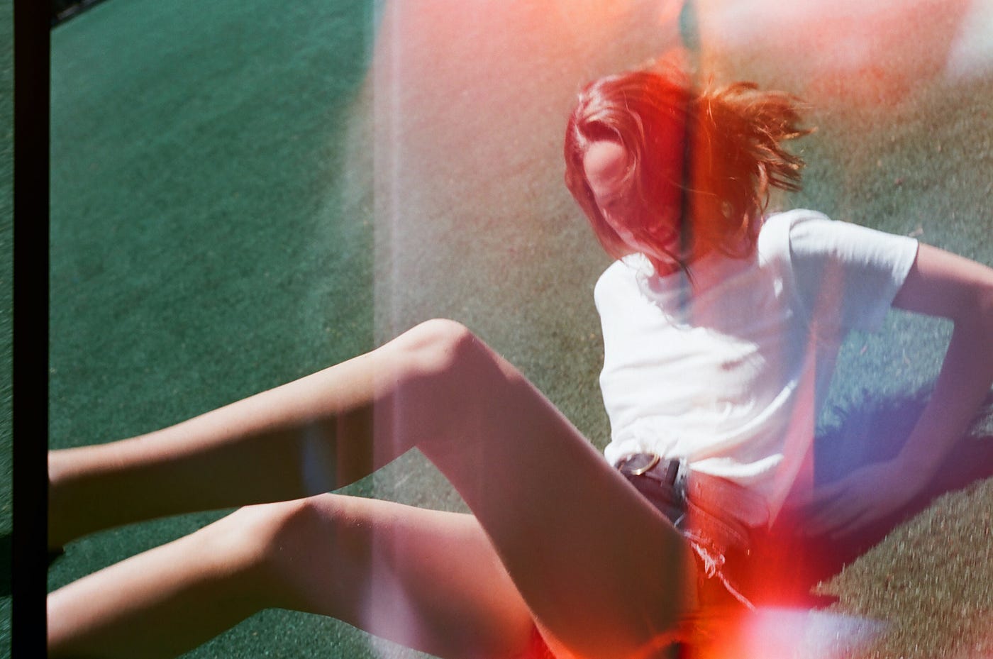 A young woman reclines on grass, face hidden by her hair. An overexposure/double-exposure effect has been added to the photo.