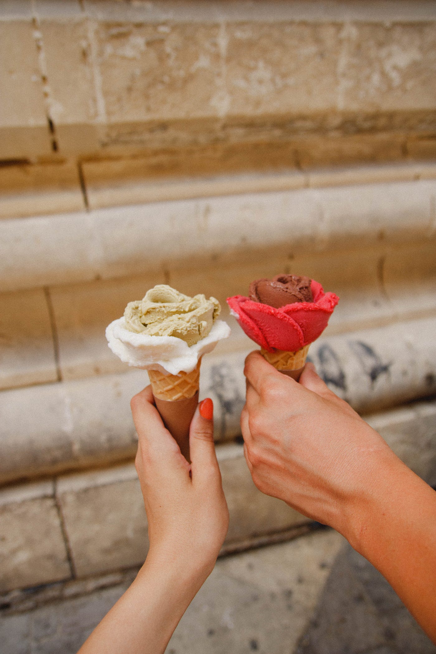Two people’s hands holding out two different ice creams. One is white on the outside and light brown on the inside, the other is dark brown on the inside and red on the outside.