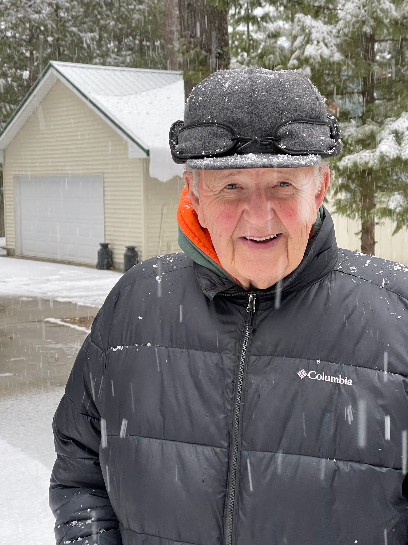 Older gentleman dressed in a winter coat and felt hat standing in front of a garage in the snow.