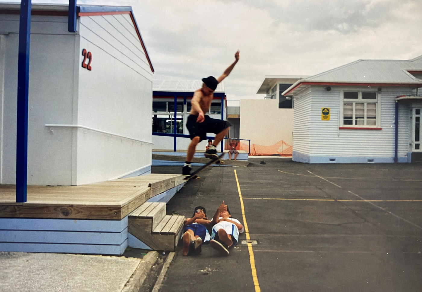 A skateboarder is captured in mid-air, he’s halfway through an olly – jumping off  two stairs and over two humans (his friends). It’s a make or break moment. He’s mid-flight directly above his two friends, who are lying down beneath him. One is throwing a shaka, the other is giving the thumbs up. Two younger kids watch the action from the distant background. They have no shoes. They wait to see if he lands it, or if he crashes into his mates.