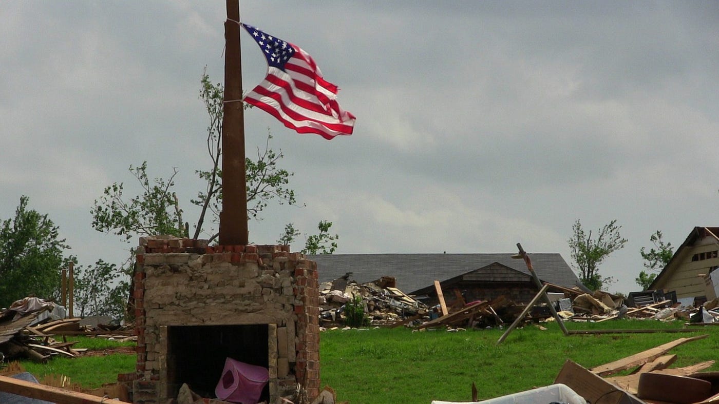 Color photo of a tragedy aftermath of a tornado, with an american flag flying in the background.