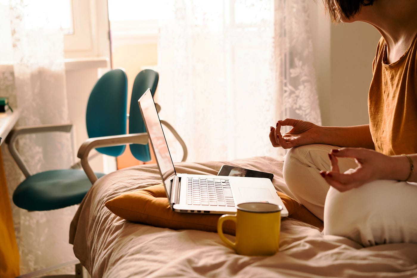 A woman in white jeans and a yellow sweater sitting on the bed in a yoga pose in front of a laptop and a cup of coffee.