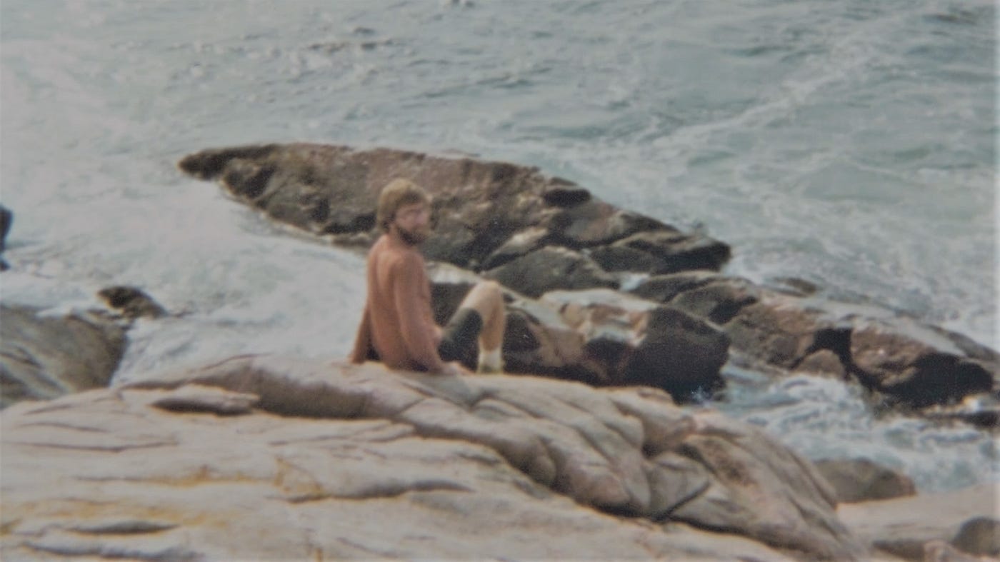 Young bearded man sitting on slanted rock jutting into the sea