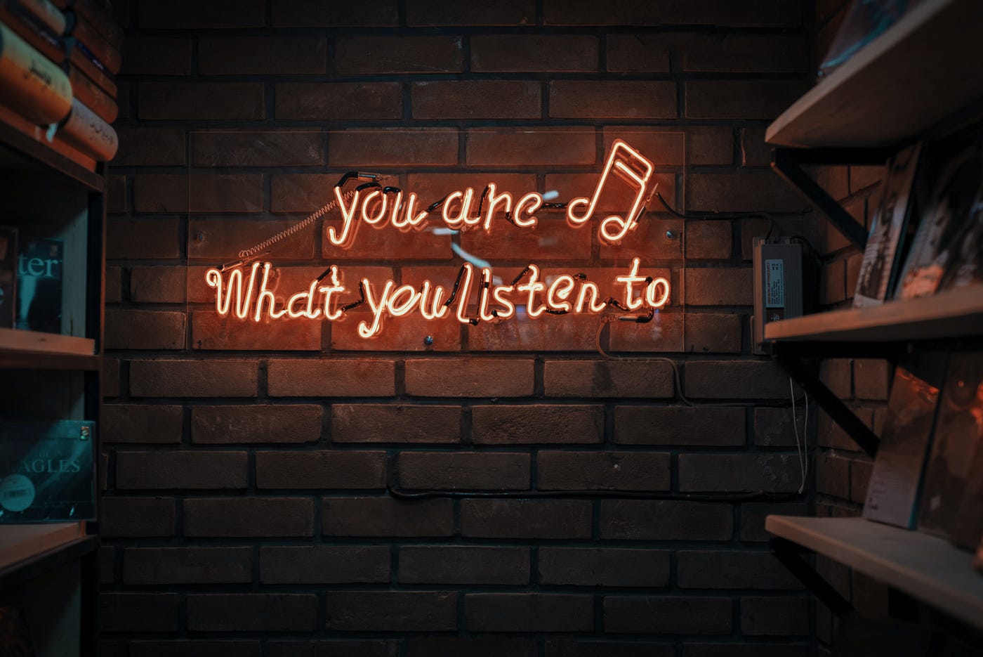photo of text ‘you are what you listen to’ in neon lights aside music notes hanging in brick wall. Photo on Dr. James Goydos 2021 article on physician satisfaction podcasts