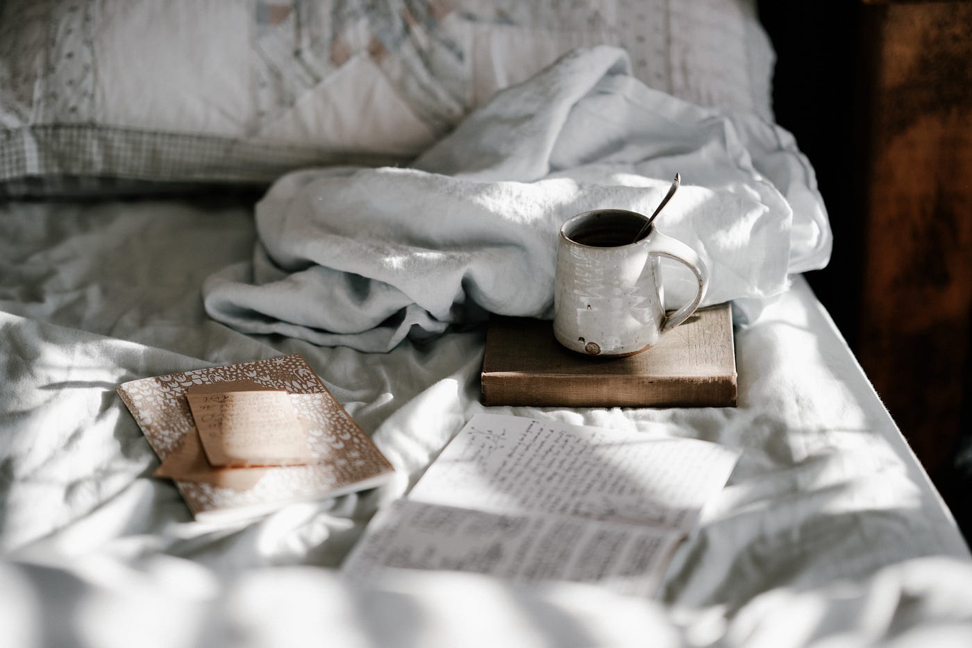 A cup of a beverage sits on a book on top of a mattress with white bedding and scattered books and journals, opened and closed lay around it.
