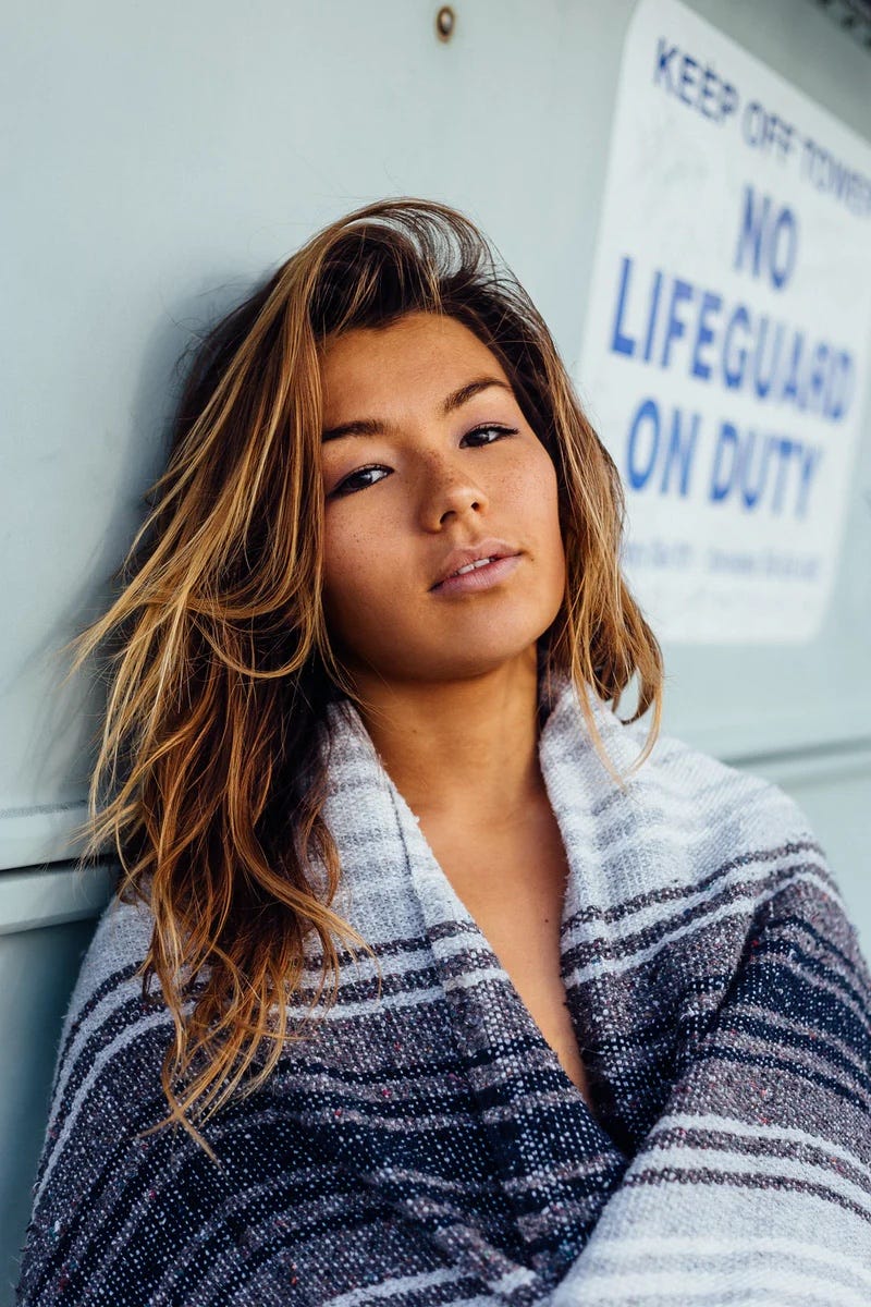 An attractive Asian woman leans against a wall, staring at us. An inscrutable expression, and maybe melasma, on her face.