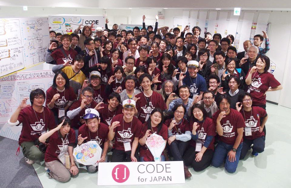 Several people pose for a group photo indoors next to a sign with the Code for Japan logo. Many people are wearing identical red t-shirts.