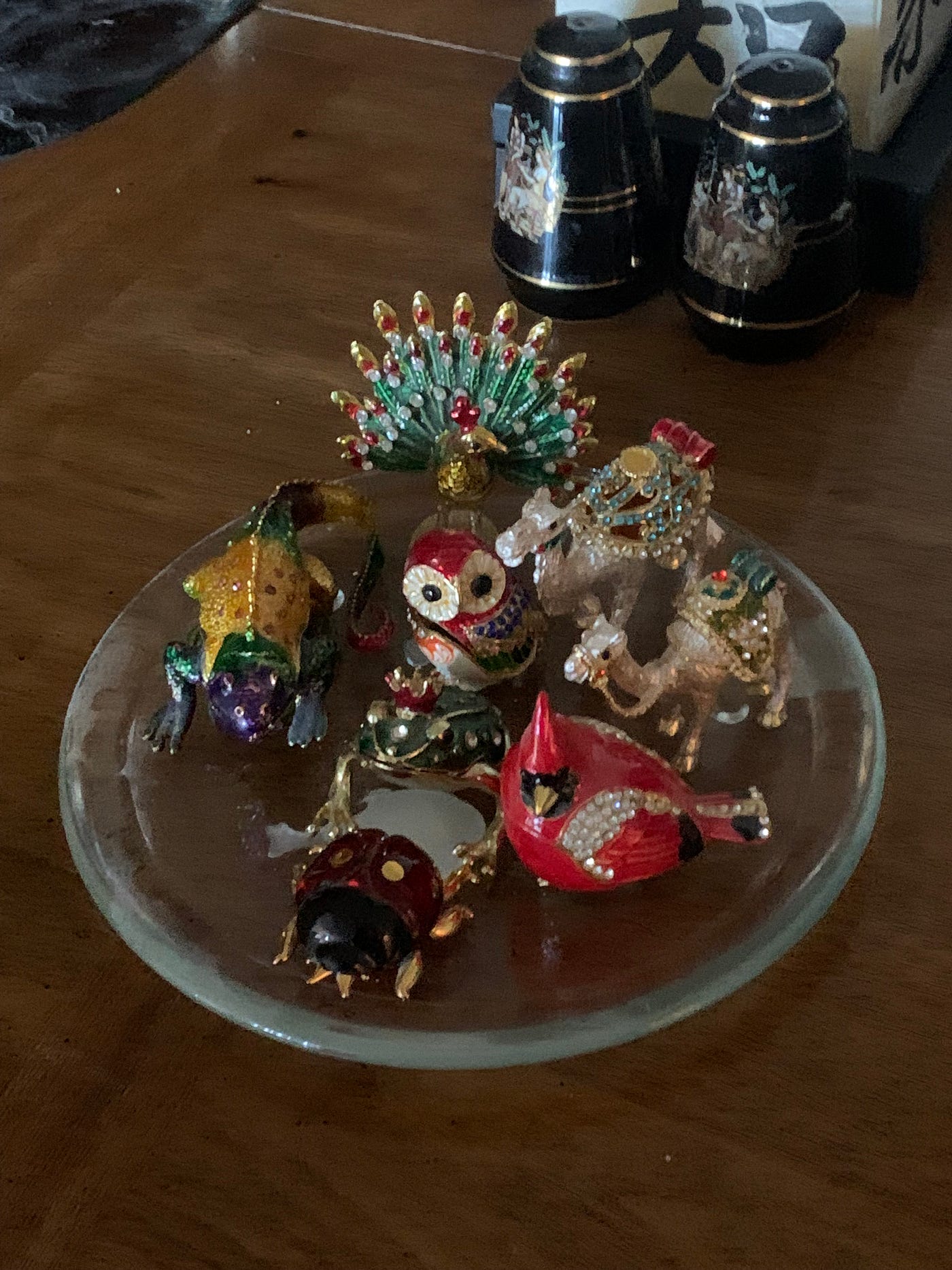 A clear white plate with a green jewelled peacock, multi-coloured lizard, two white camels, a green frog, a red robin and a red, black and gold lady bug ring holder on display on my mahogany coffee table.
