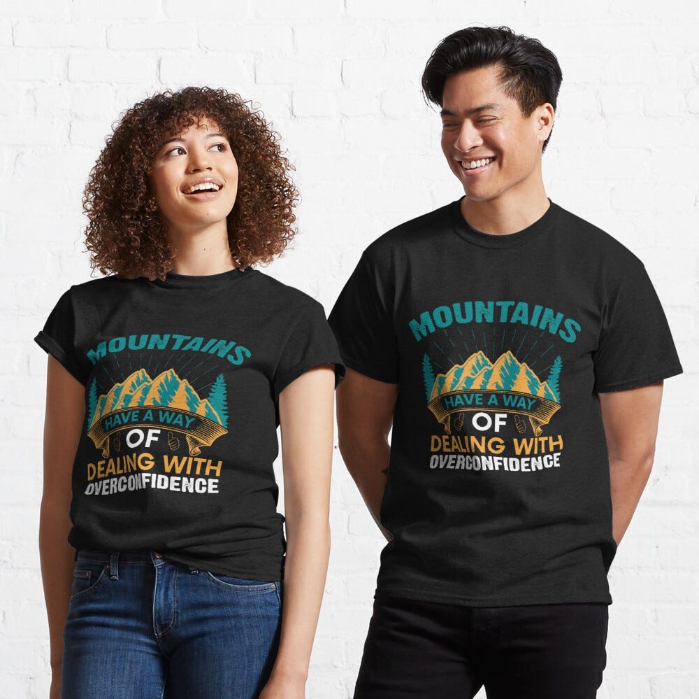 Man and Woman Wearing a black classic t-shirt with amazing illustration and text saying “mountains have a way of dealing with overconfidence”.