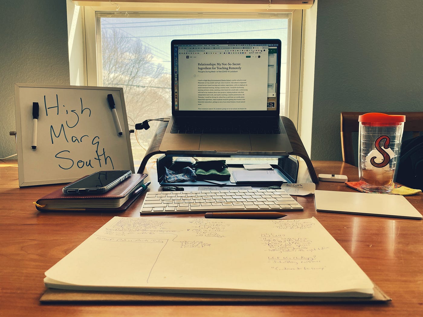 A laptop computer and various desk accessories laid out on a dining room table in front of a window