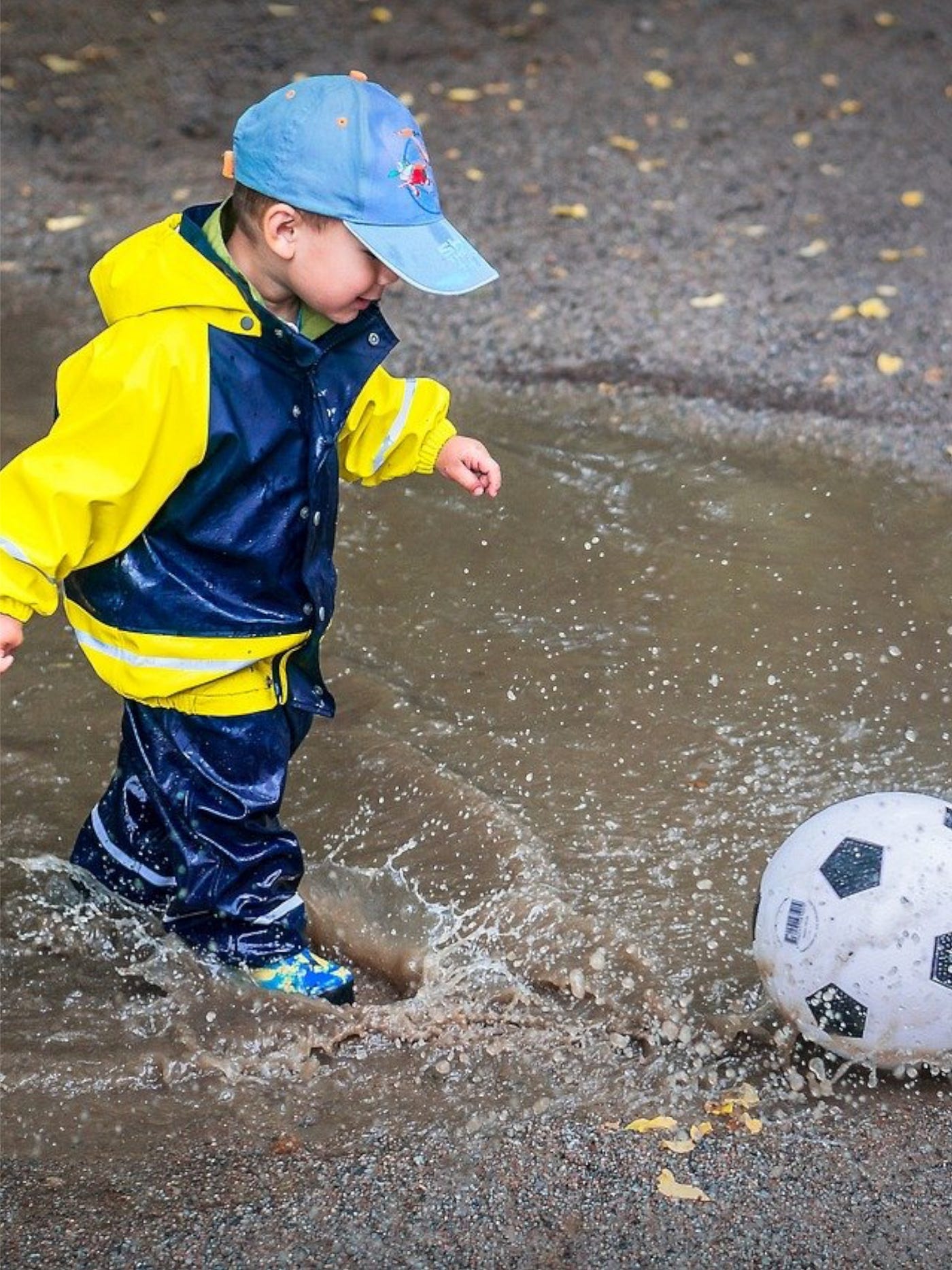 Color photo of a young boy stomping through a puddle, kicking a soccer ball.