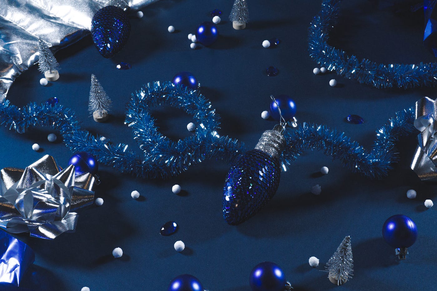 A collection of blue and silver ornaments, gift bows, pompoms and strings of tinsel lay arranged on a flat blue surface.