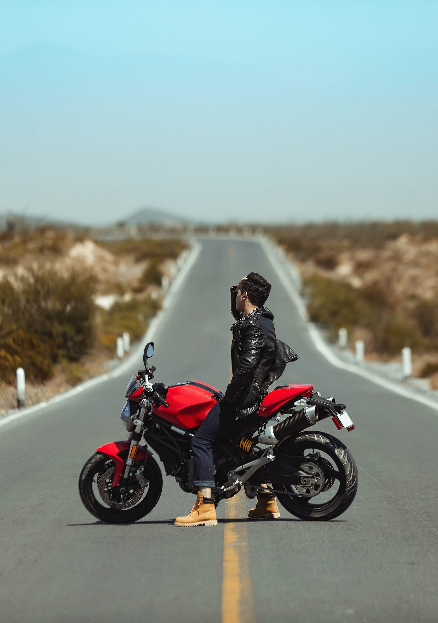 A young man rakes his fingers through his hair while sitting on a motorcycle on a long empty road