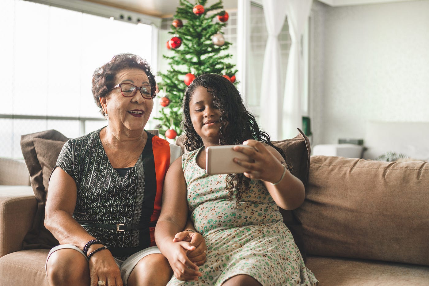 A young girl and her grandmother take a selfie on their couch in front of a Christmas tree.