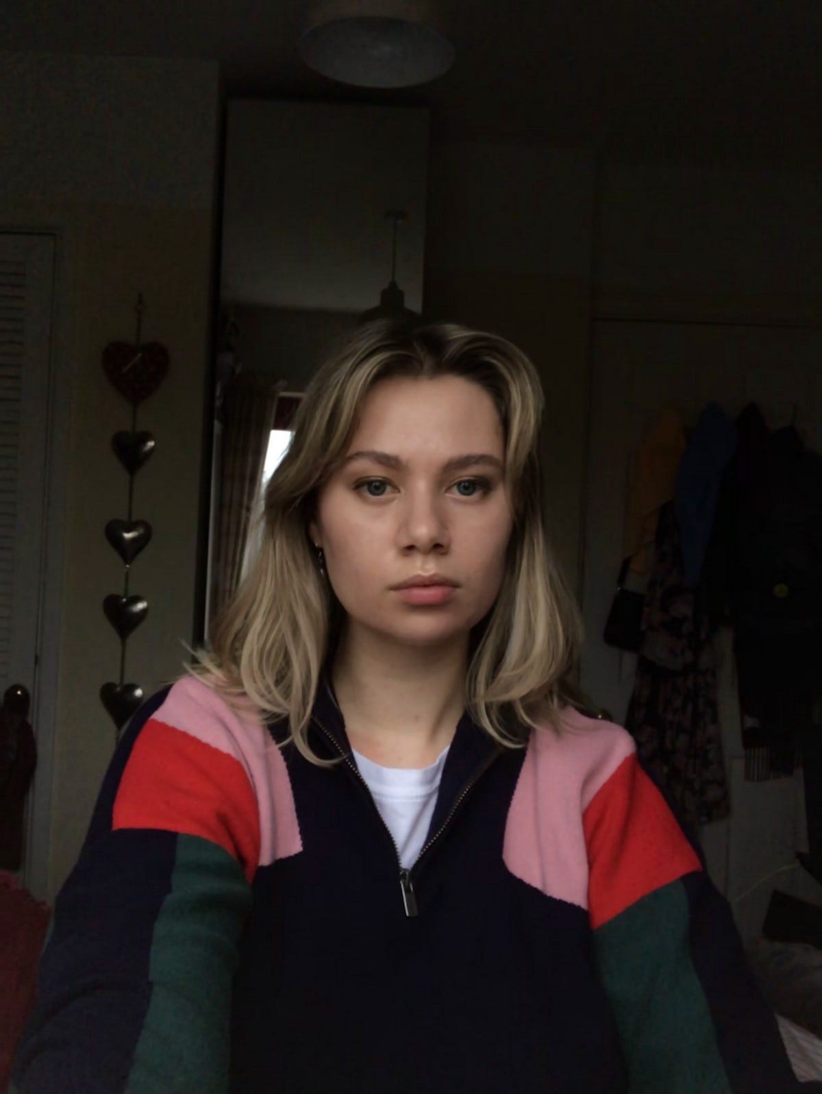 Eden sat on her bed with dark blonde hair, wearing a navy blue jumper with pink red and green sleeves. She is straight faced and looking into the camera.