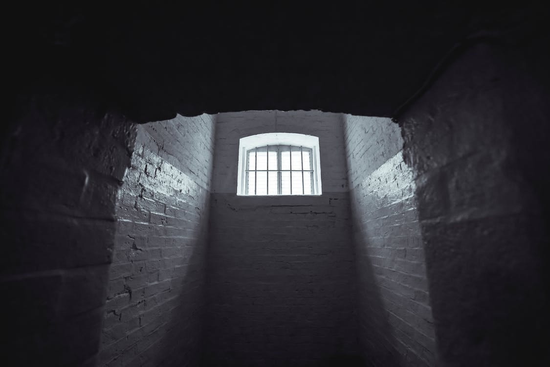 A dark prison cell with light shining through a grated window.
