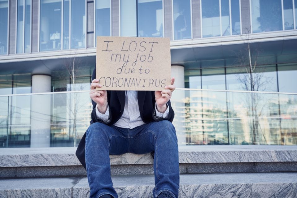White male (face not seen behind the cardboard sign) in jeans and a sport coat sitting in front an office building. He’s holding a crudely-made sign that reads “I lost my job due to Coronavirus”