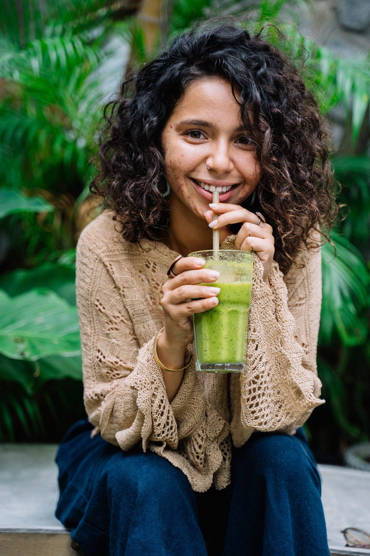 A woman in a brown sweater and jeans drinks a green smoothie.