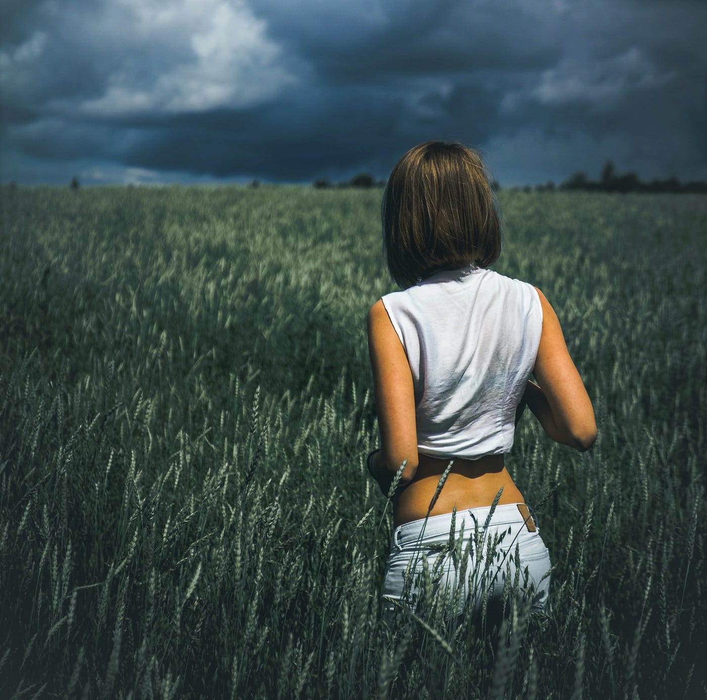 back view of girl with brown hair wearing white top and pants standing in grassy field looking at stormy sky