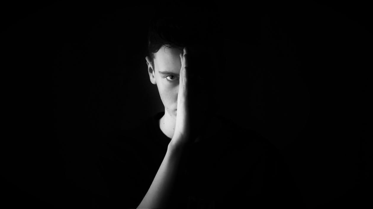 A man against a black background holding up a hand that is dividing his face. The right side of his face is blacked out.