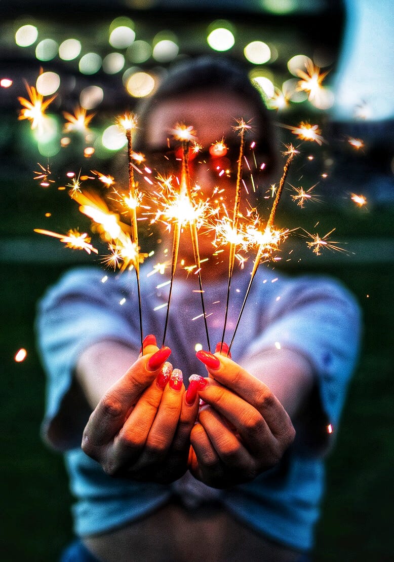 A woman with long red painted nails is holding up five lit sparklers
