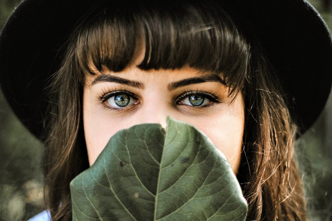 girl with brown hair and green eyes wearing dark hat holding leaf in front of her face