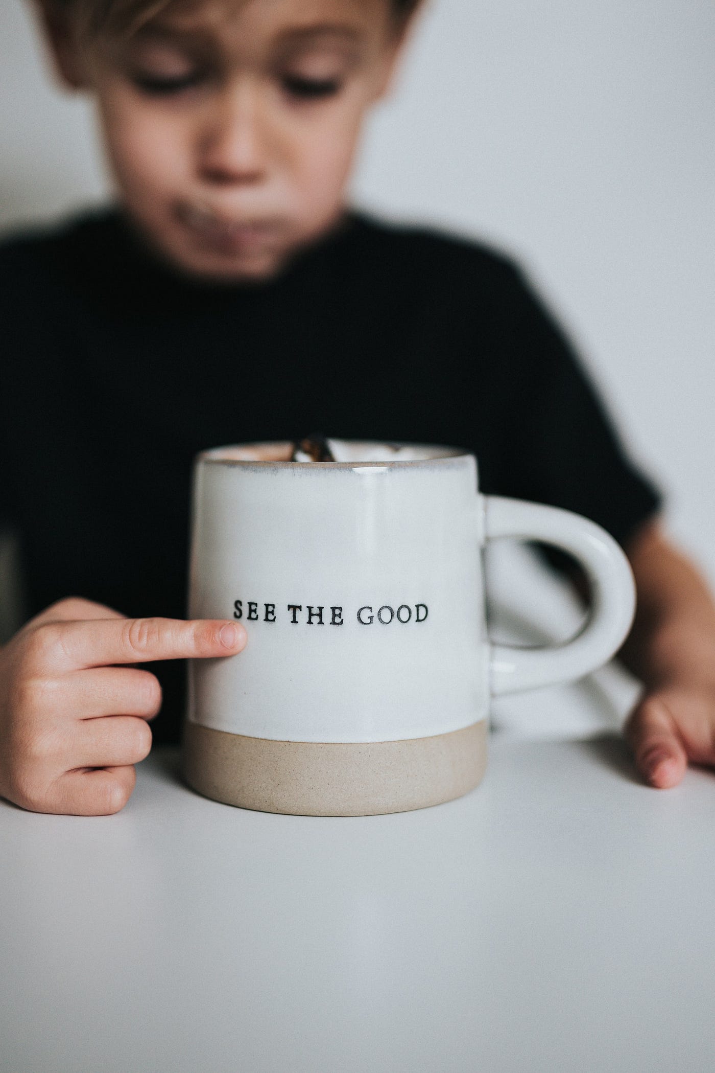 A young boy sits at a table pointing at a coffee cup that reads: See the Good (in everybody)