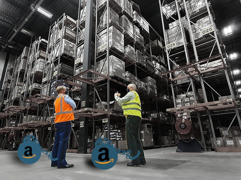 Two warehouse workers surrounded by high shelves laden with merchandise; each one has a leg shackled to a ball-and-chain bearing the Amazon ‘A’ logo.