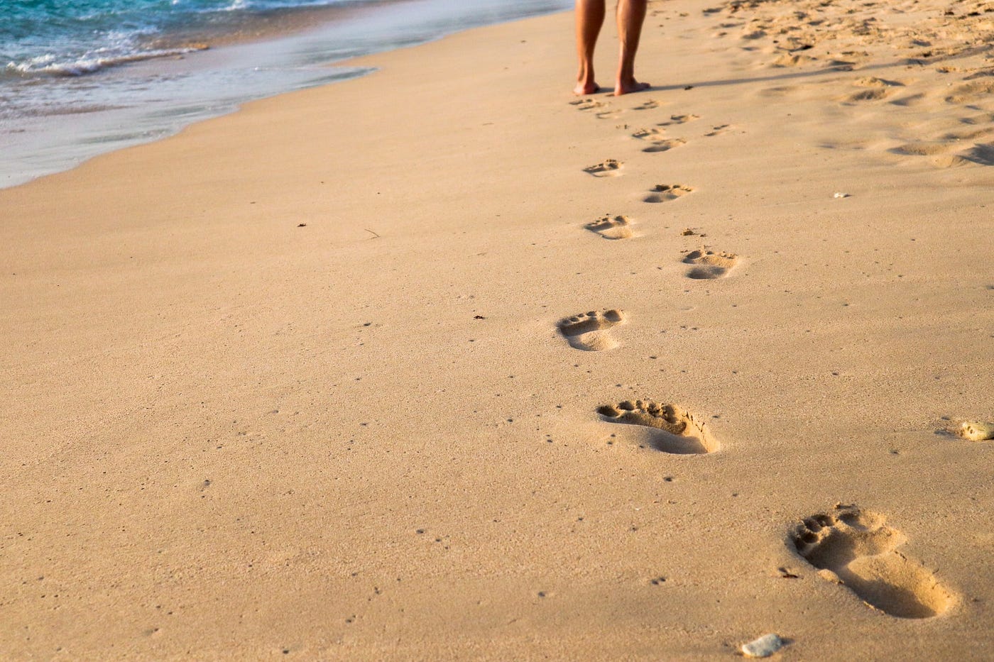 Barefoot footsteps in sand next to water.