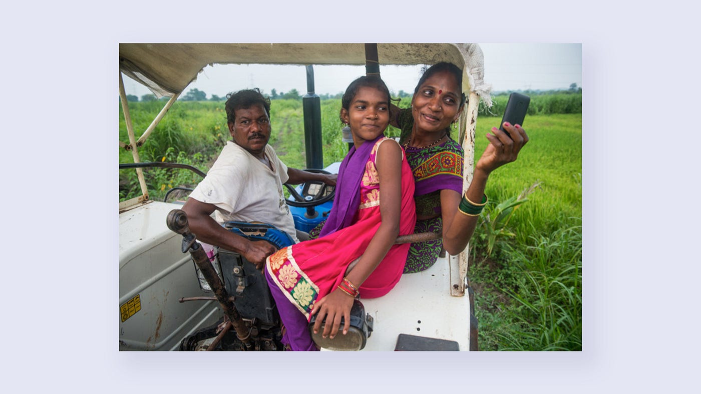 Three people ride a utility vehicle in a field while one holds a phone, holding it so the camera can view all three people.